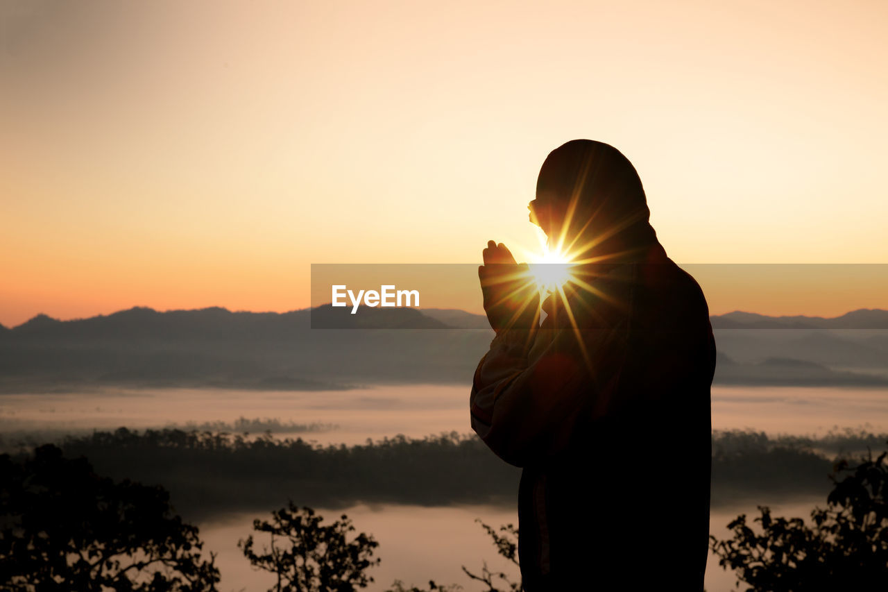 Side view of man praying against sky during sunset