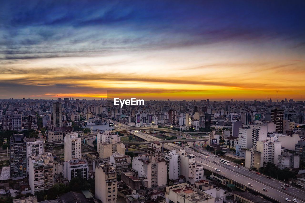 HIGH ANGLE VIEW OF MODERN BUILDINGS IN CITY AGAINST SKY DURING SUNSET