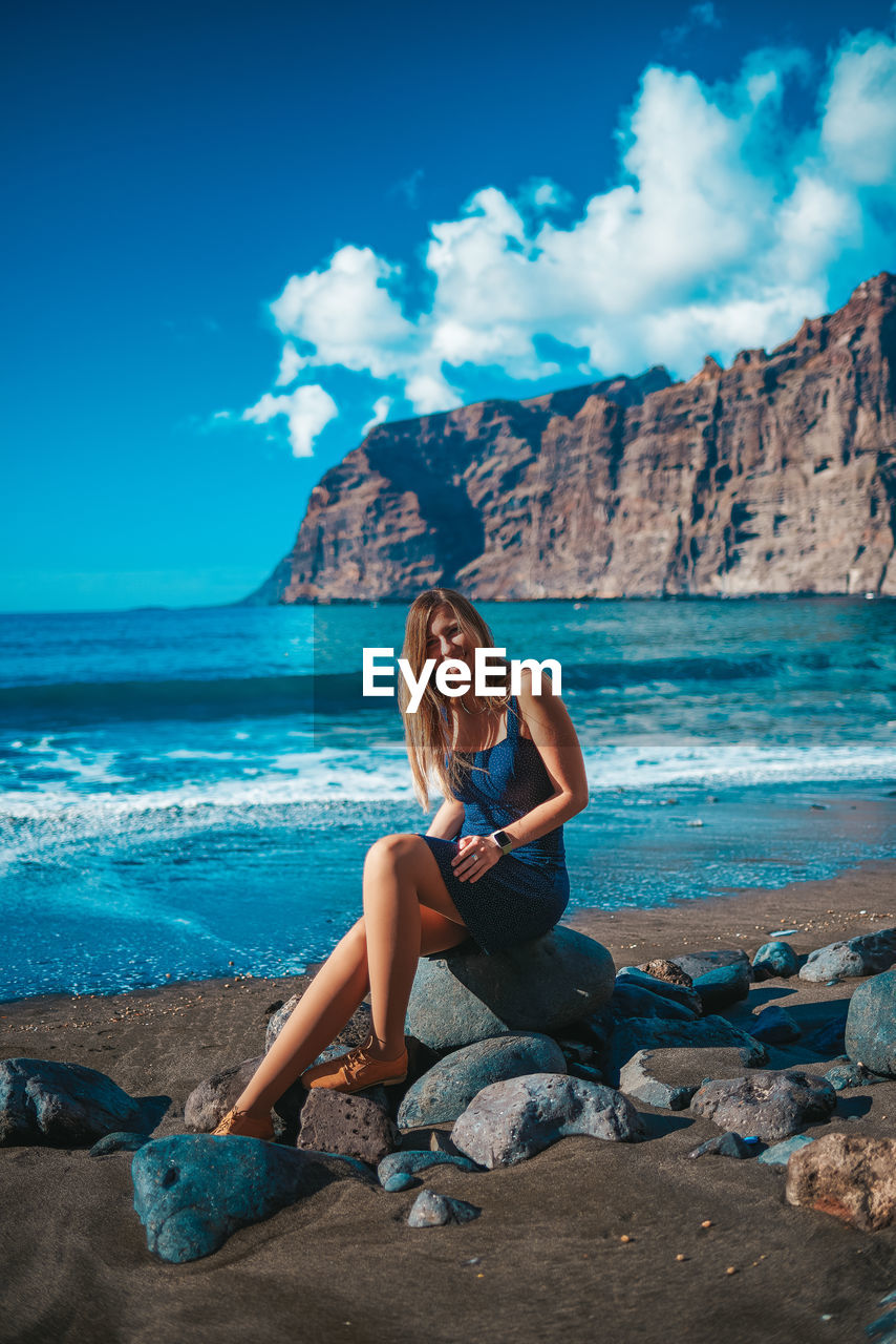 FULL LENGTH OF YOUNG WOMAN SITTING ON ROCK AT BEACH