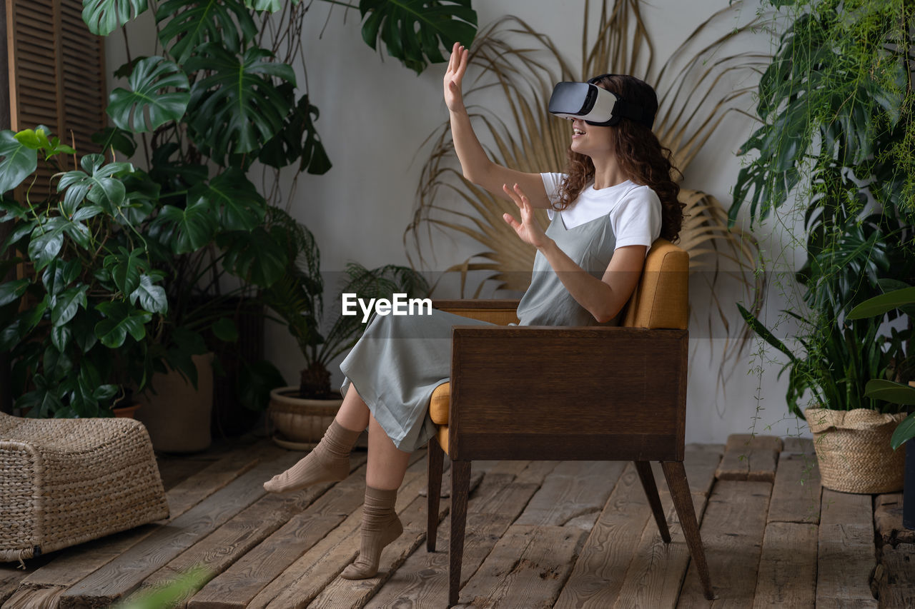 Young amazed woman in vr headset resting at indoor greenhouse full of tropical and exotic plants