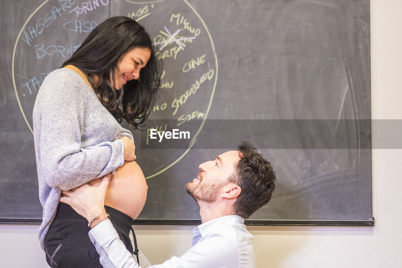 Side view of man kneeling in front of pregnant girlfriend against blackboard