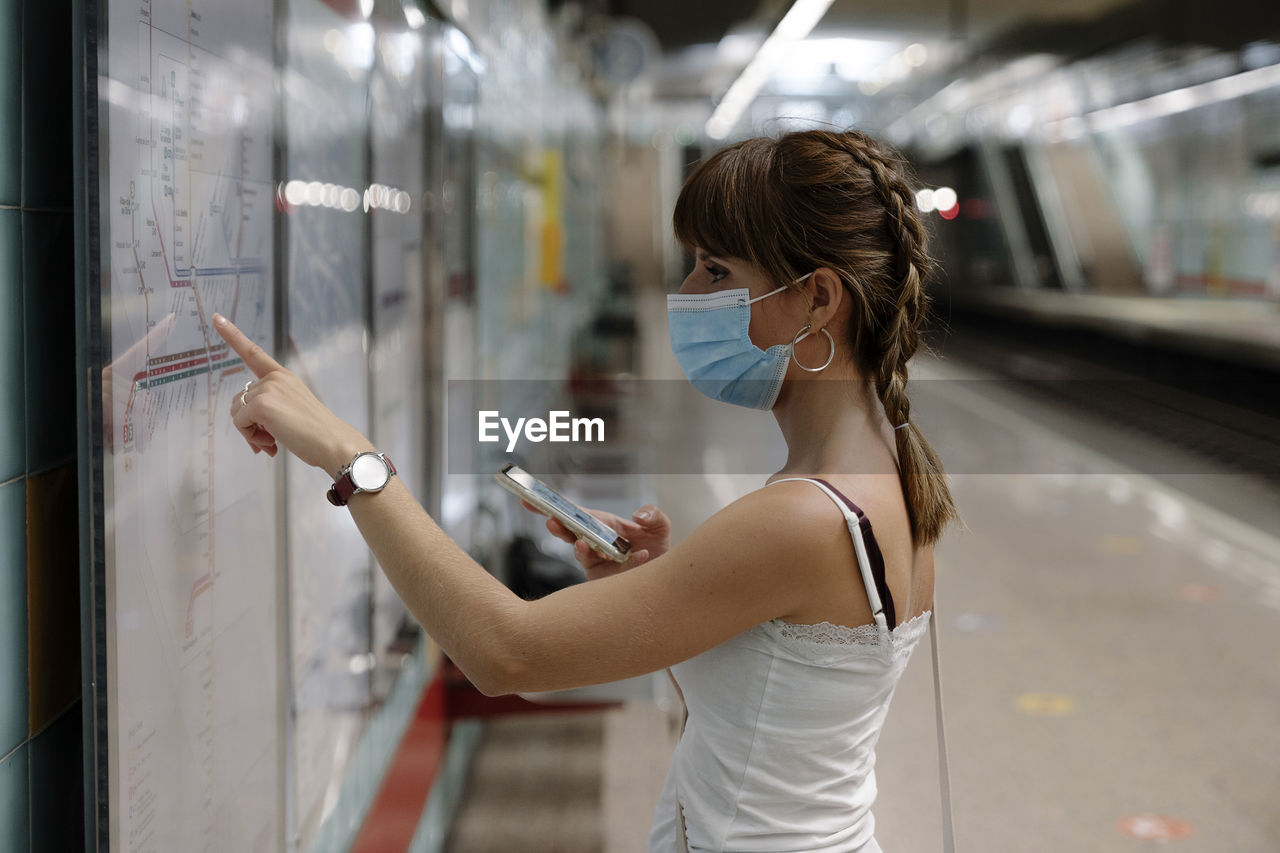 Woman with protective face mask looking at rout map of train on platform during pandemic