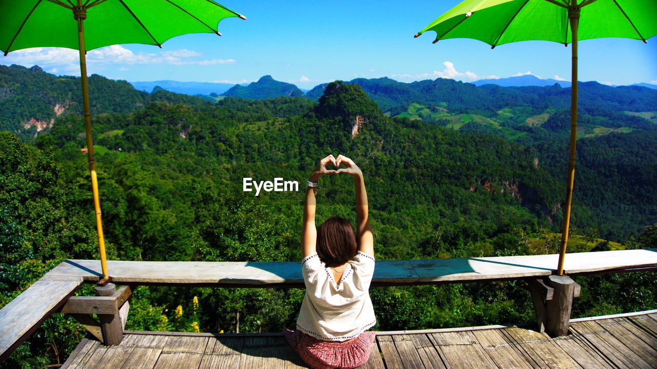 Woman making heart shape on footbridge against blue sky