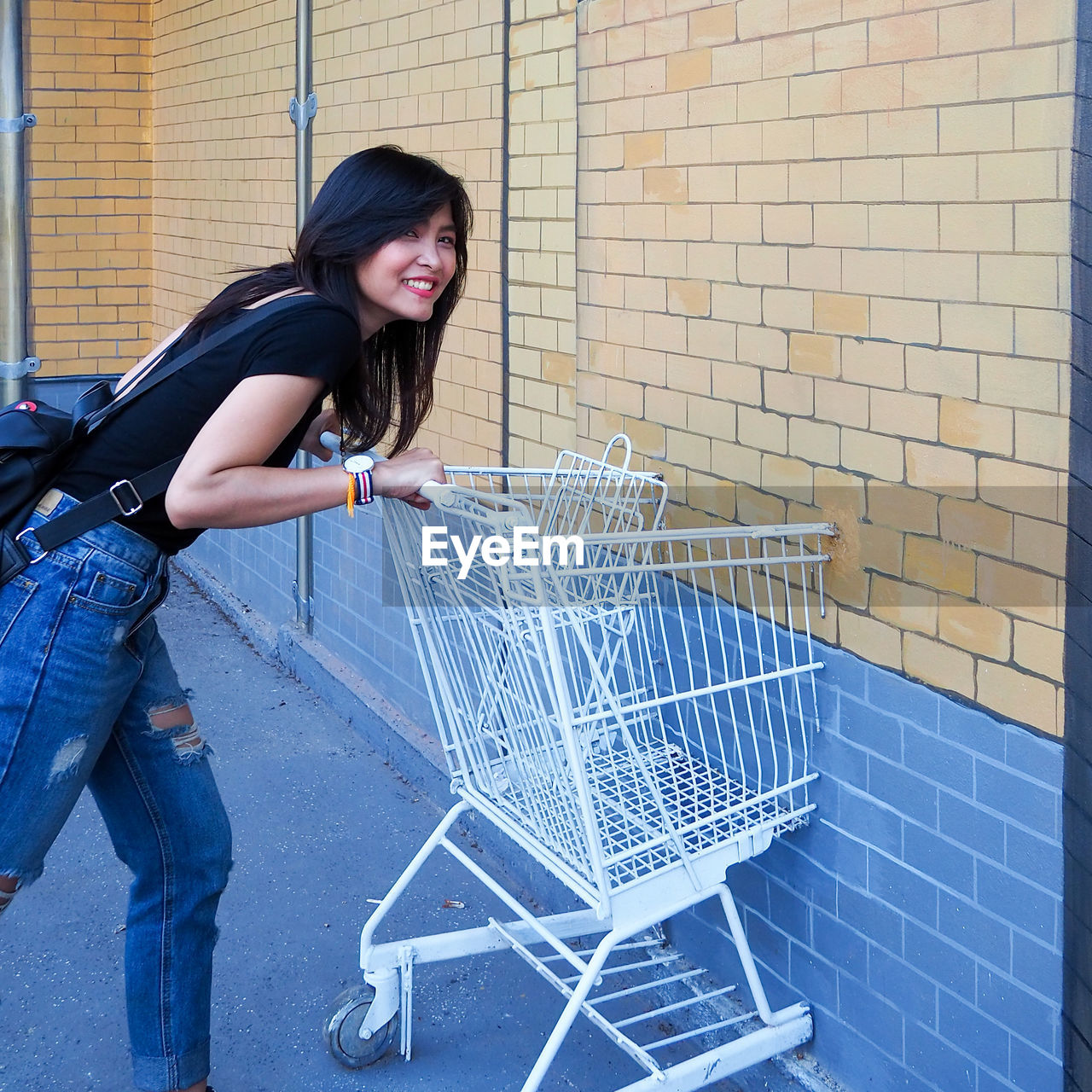 Side view portrait of smiling woman holding shopping cart by brick wall