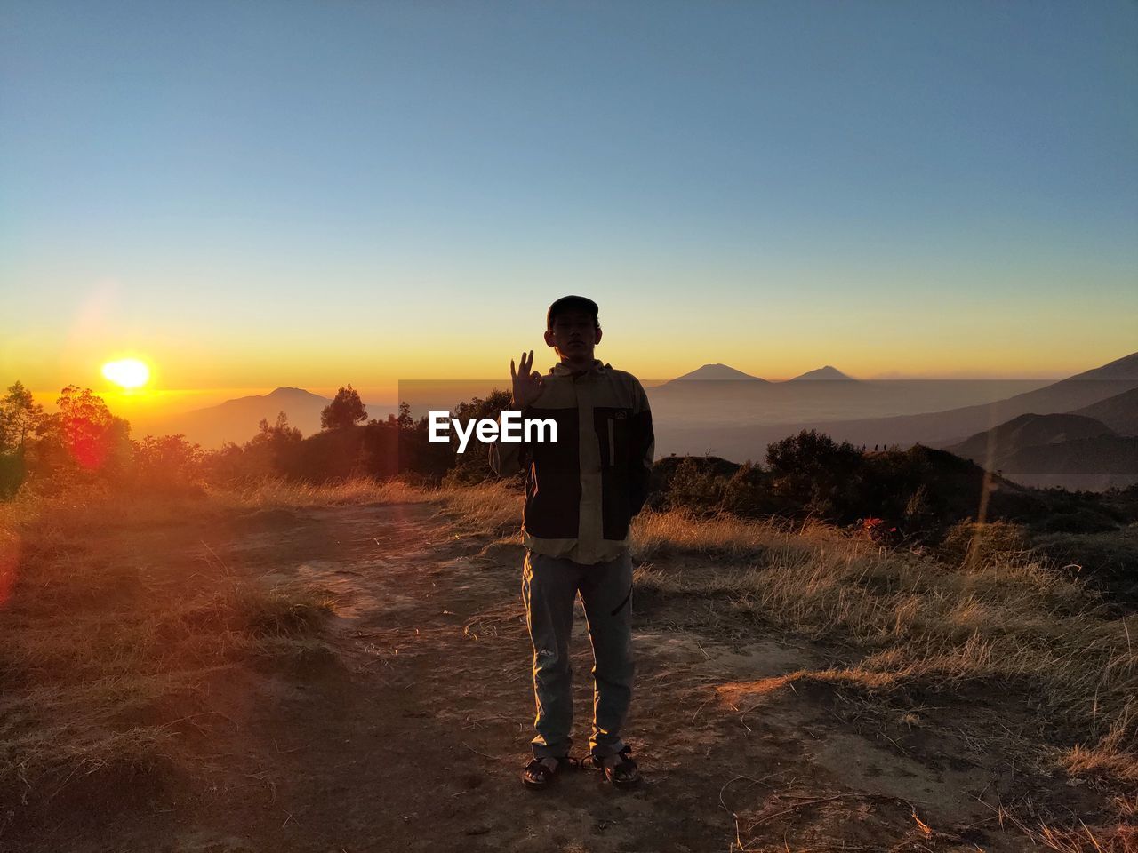 REAR VIEW OF MAN STANDING ON LAND AGAINST SKY DURING SUNSET