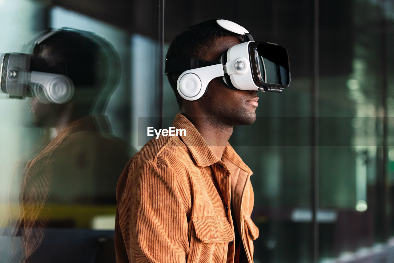 Positive african american male in modern vr goggles standing near glass wall while exploring virtual reality on street of city