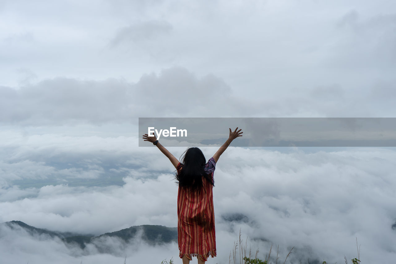 Rear view of woman with arms raised standing against cloudy sky