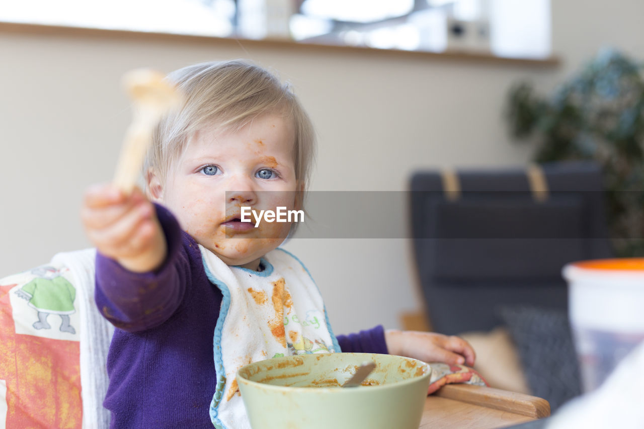 Portrait of cute baby girl with messy face sitting with food bowl at home