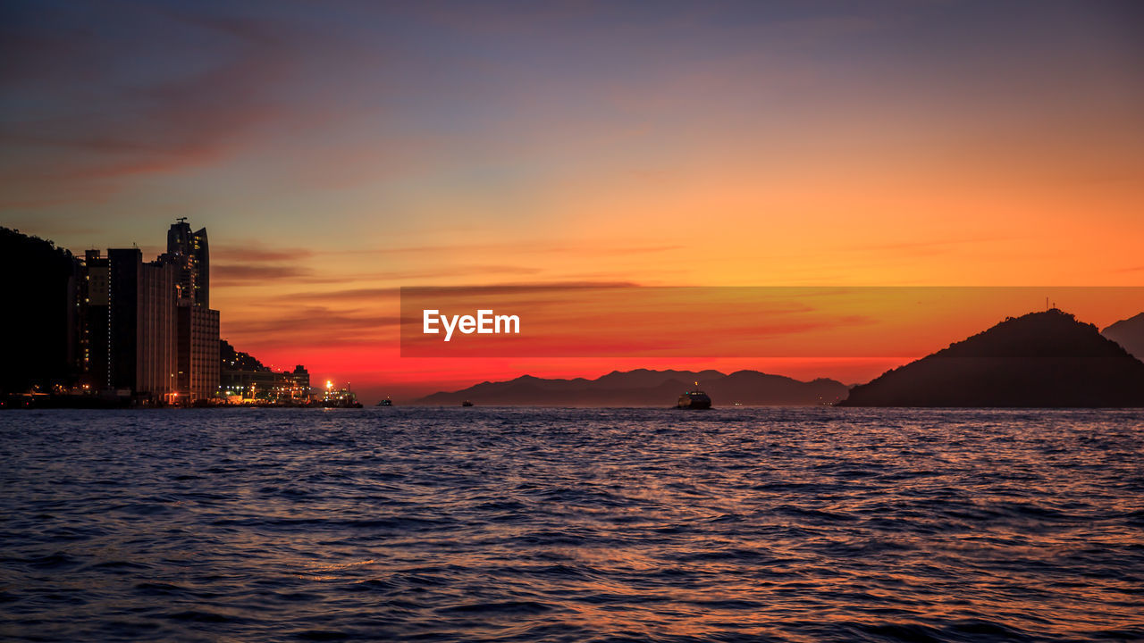 Scenic view of sea by silhouette buildings against sky during sunset