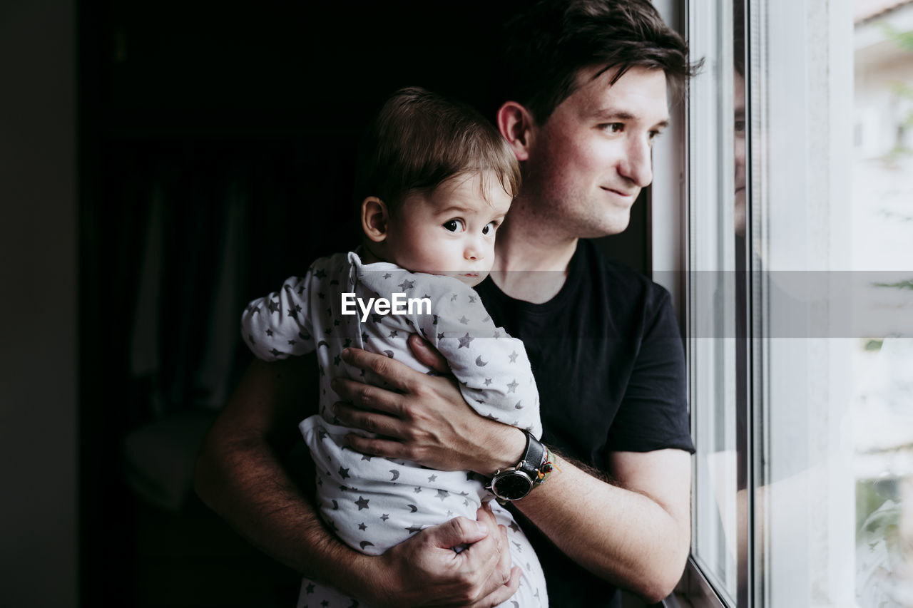 Smiling father carrying son while standing looking through window at home