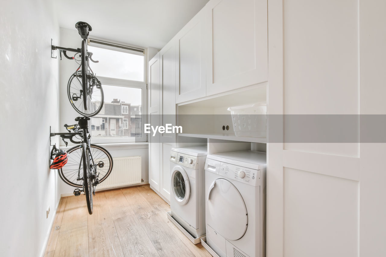 Modern white electric washing and dryer machines placed near white cabinets and basket in light laundry room with window and bicycle