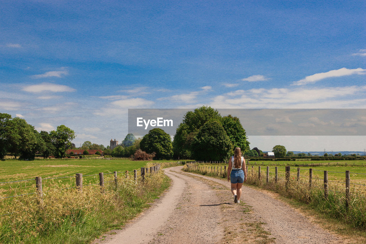 Rear view of woman walking on track through field towards trees with a blue sky.