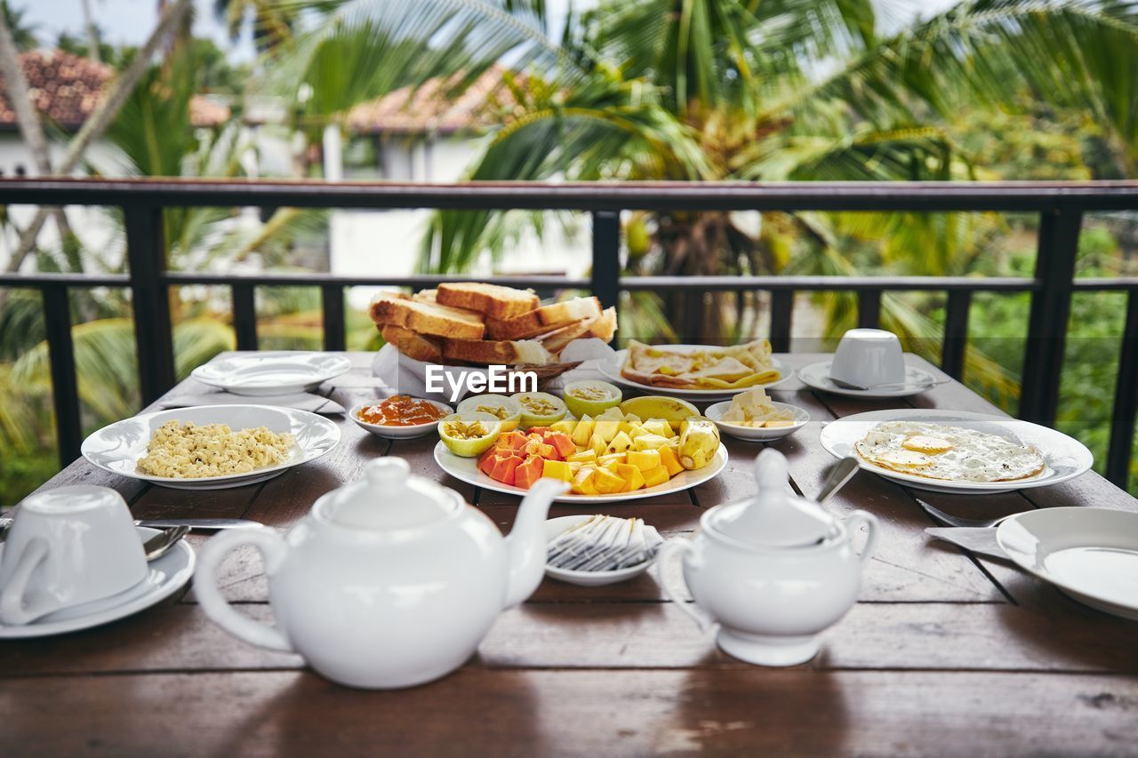 Breakfast ready to eat. eggs, tropical fruit, toasts and tea pot on wooden table against palm tree.