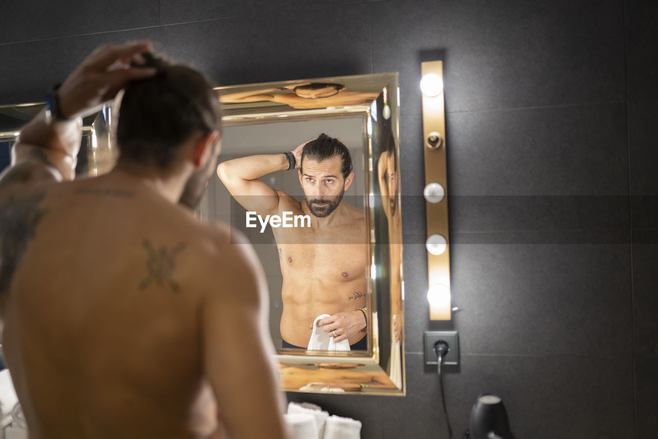 Shirtless tattooed male looking at mirror and doing ponytail in illuminated contemporary bathroom