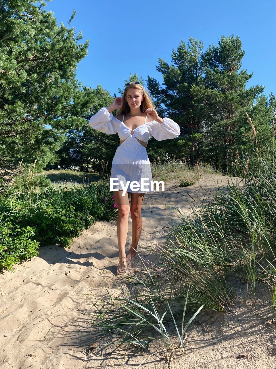 one person, plant, women, full length, nature, adult, young adult, vacation, leisure activity, tree, casual clothing, front view, happiness, smiling, standing, day, emotion, lifestyles, long hair, hairstyle, summer, clothing, sunlight, sky, portrait, land, blond hair, outdoors, fashion, holiday, trip, female, barefoot, shorts, carefree, looking at camera, enjoyment, person, landscape, blue, sunny, cheerful, human leg, environment, beauty in nature, rural scene, spring, grass, footwear, relaxation, growth