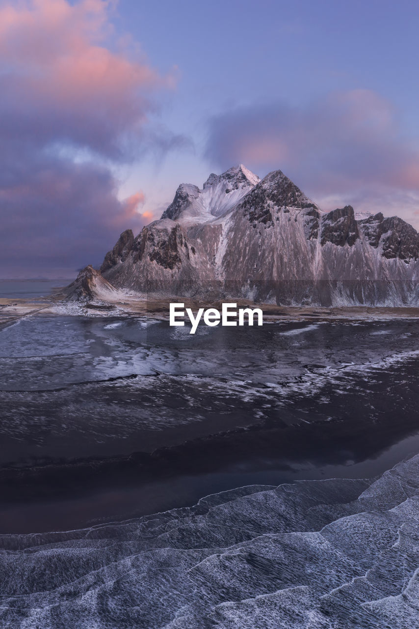 Drone view of calm frozen endless sea surrounding high rocky snowy vestrahorn mountain during picturesque sunset in stockness beach, iceland