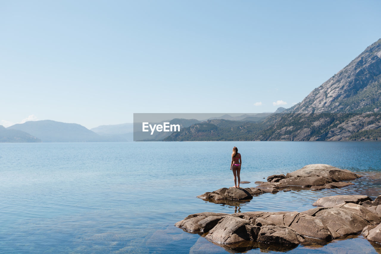 Rear view of woman in bikini standing at lakeshore against clear sky during sunny day