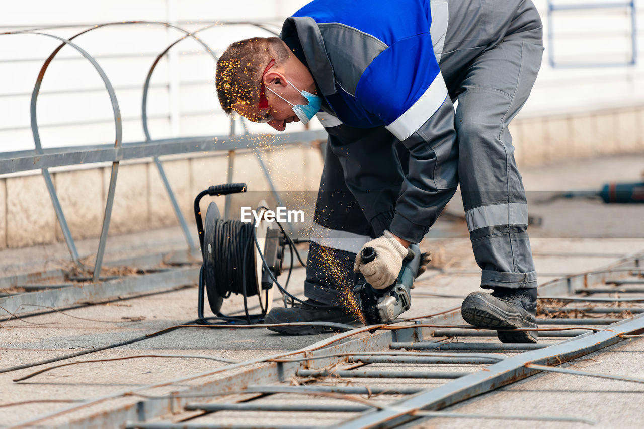 Builder in overalls leans over and cuts off metal sheet with angle grinder and sparks fly. working