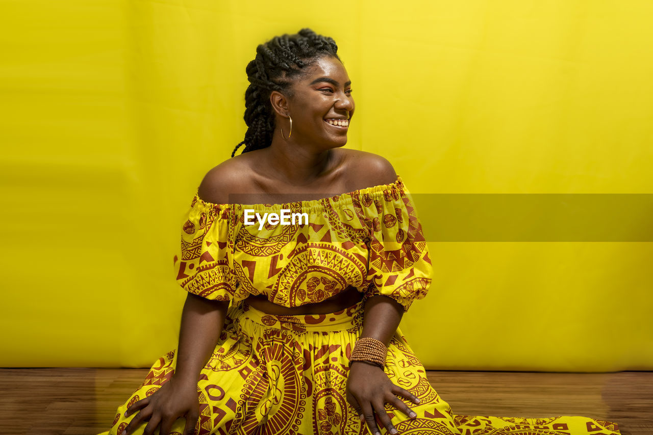 Young beautiful woman sitting on wooden floor, wearing colorful dress. against yellow background.