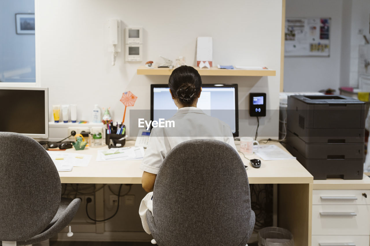 Rear view of female doctor working on desktop computer while sitting on chair at clinic