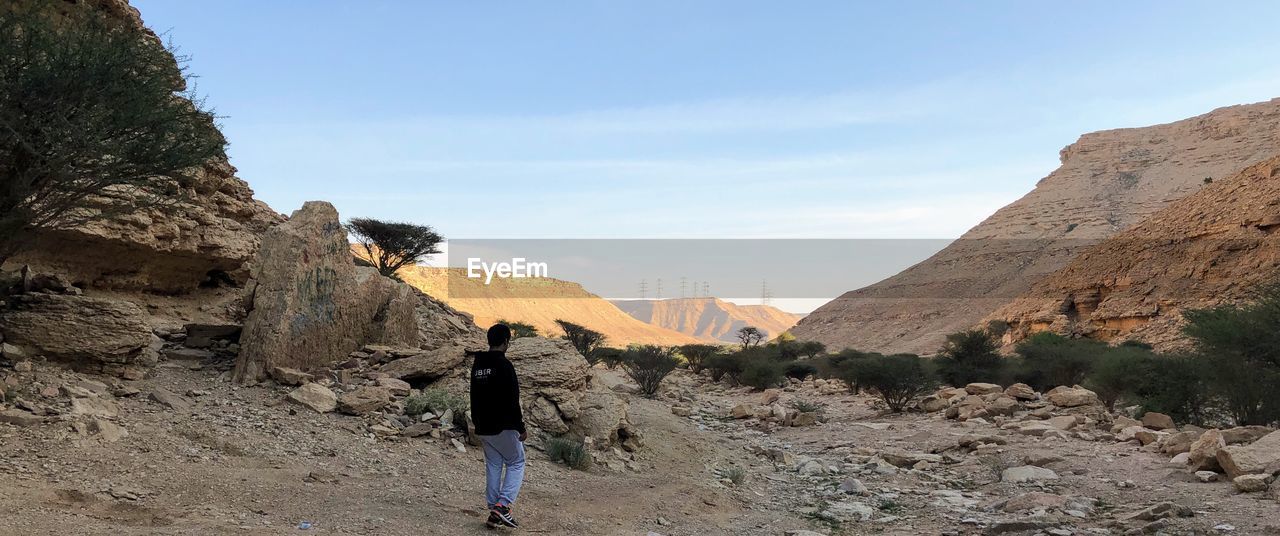 REAR VIEW OF MAN STANDING ON ROCKS AGAINST SKY