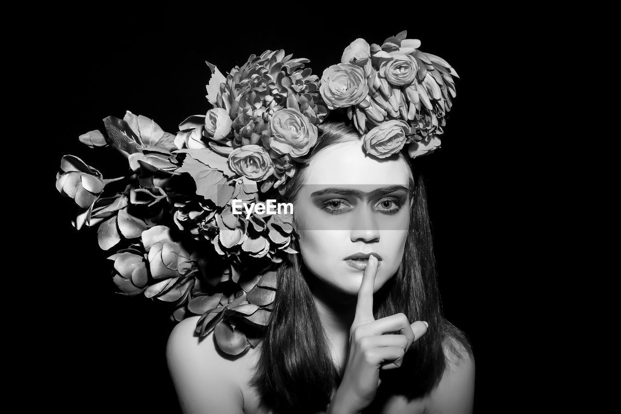 Portrait of fashion model wearing flowers with finger on lips against black background