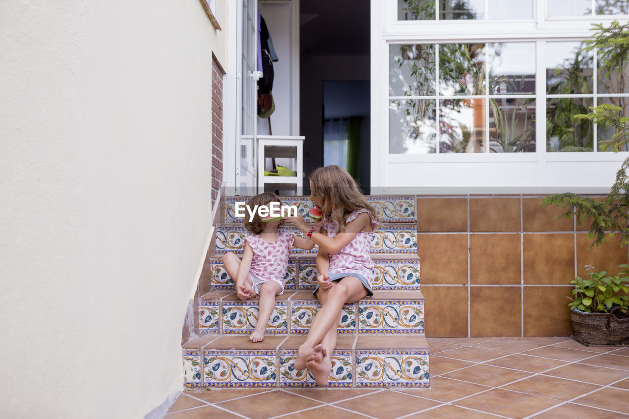 Sisters feeding watermelon to each other against house