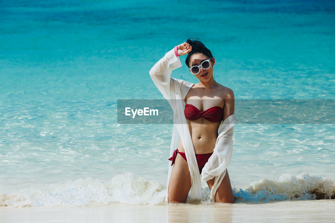 Portrait of young woman wearing bikini and sunglasses kneeling on shore at beach