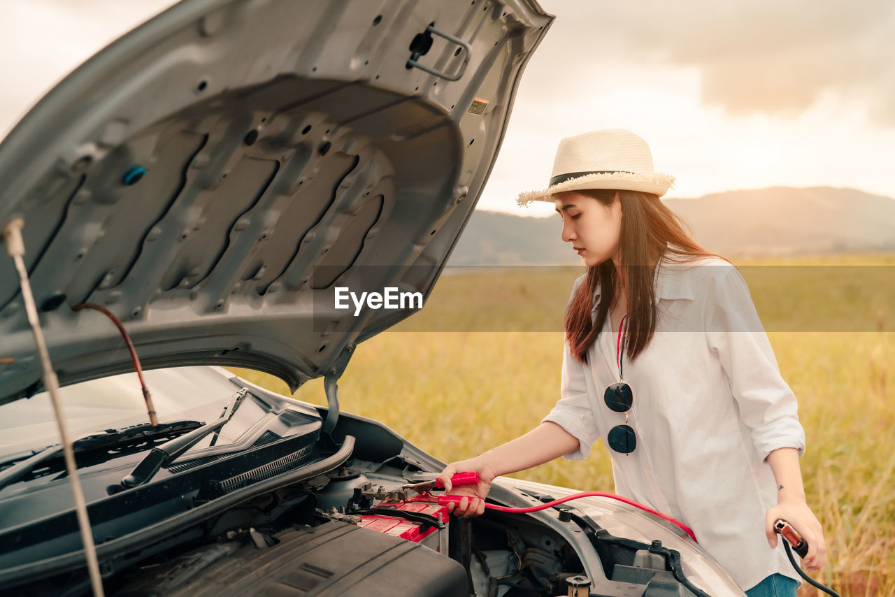 Young woman examining car on land against sky