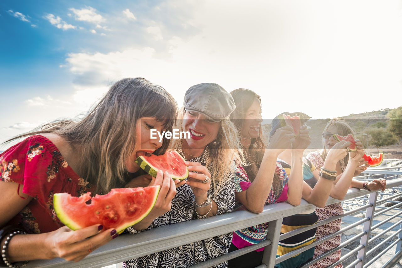 Happy friends eating watermelons by railing against sky