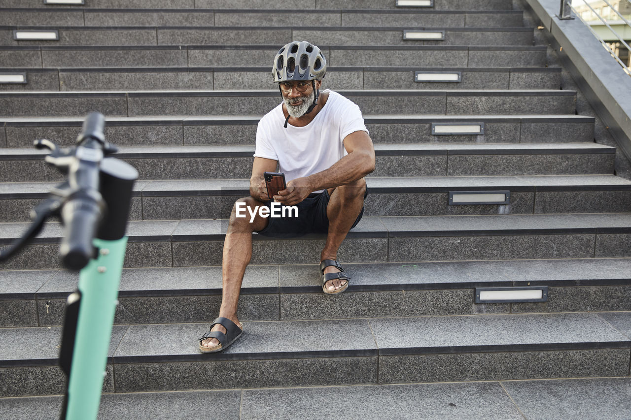 Smiling mature man with e-scooter sitting on stairs using cell phone