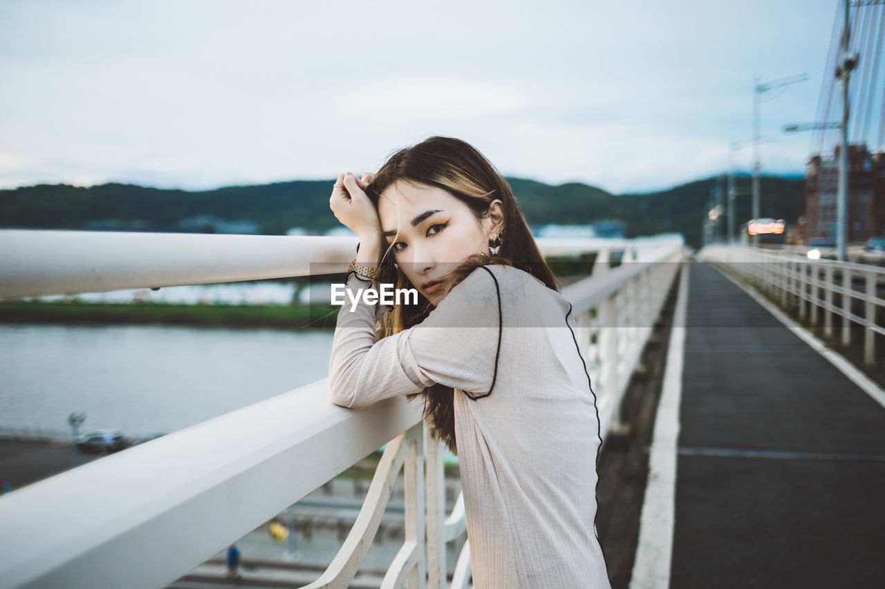 PORTRAIT OF YOUNG WOMAN STANDING ON RAILING AGAINST WATER