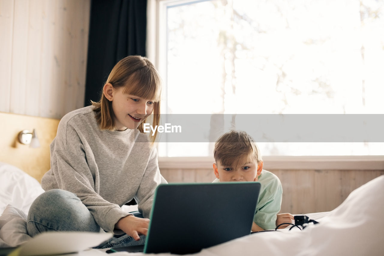 Smiling girl with brother using laptop in bedroom at home