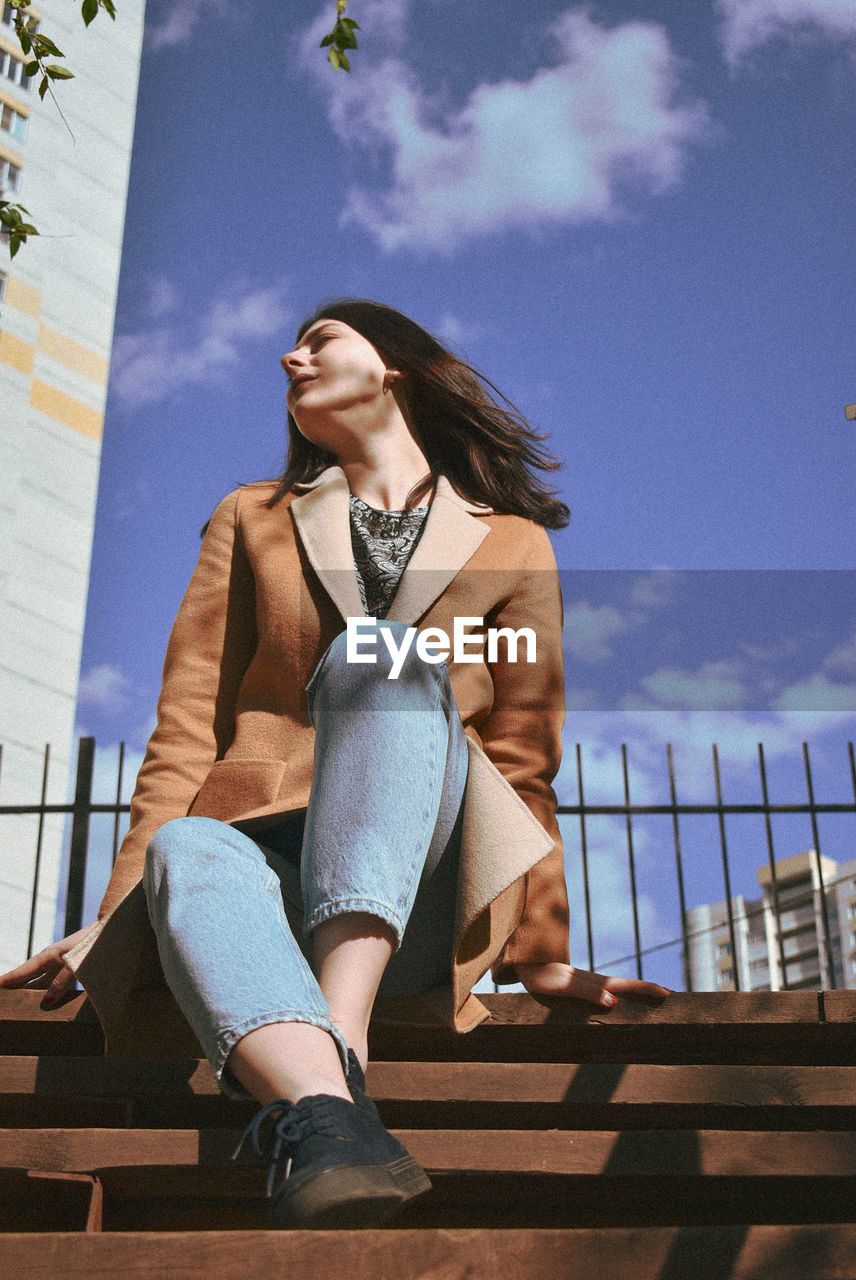 LOW ANGLE VIEW OF WOMAN LOOKING AWAY WHILE SITTING ON RAILING