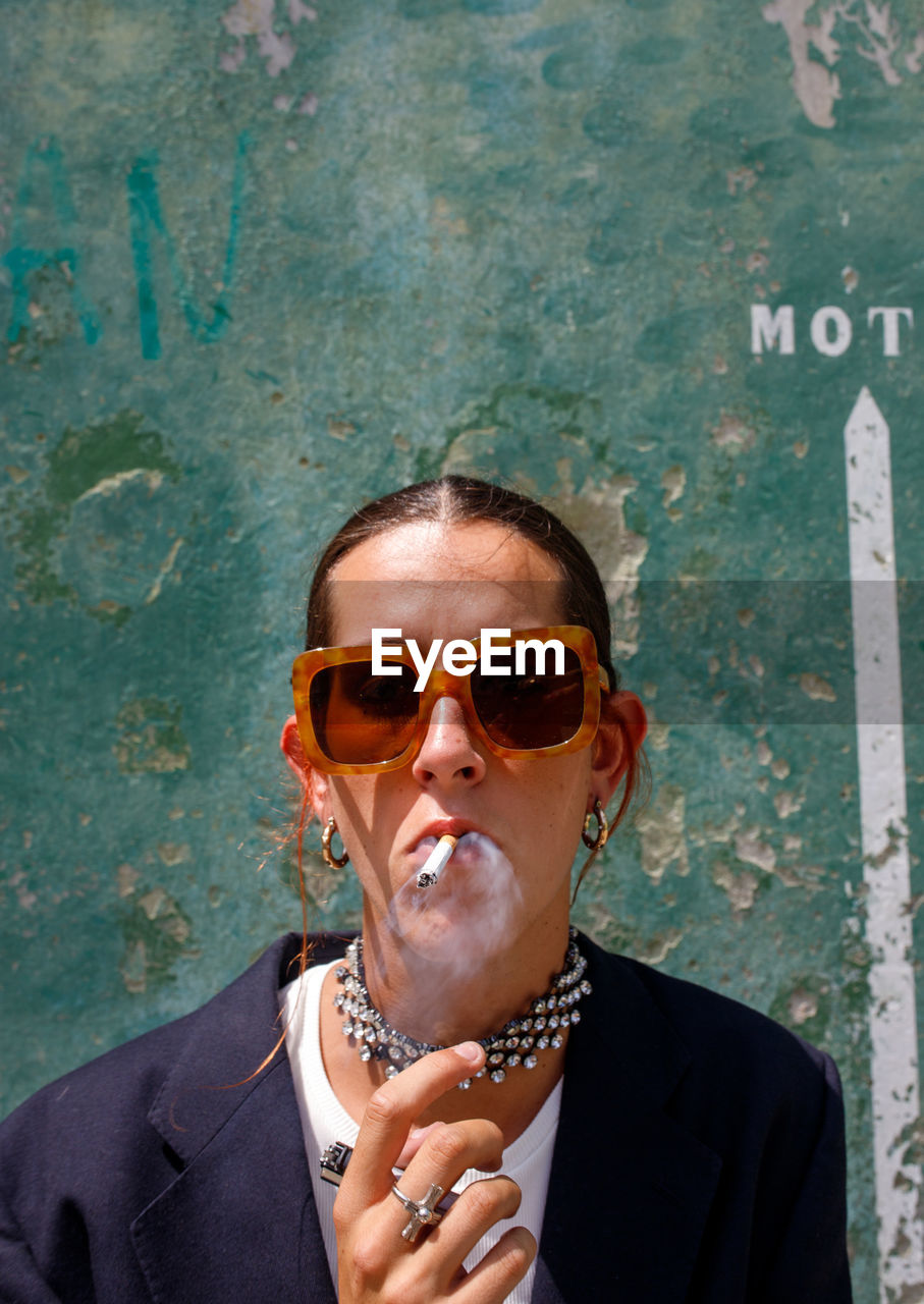 Self assured adult female with dark hair in stylish outfit and sunglasses smoking cigarette and looking at camera on street near shabby wall on sunny day