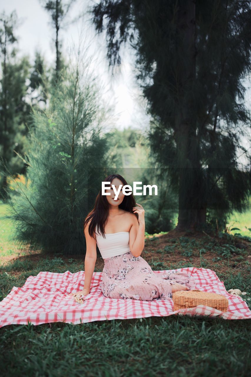 Beautiful young woman sitting on picnic blanket against trees in forest