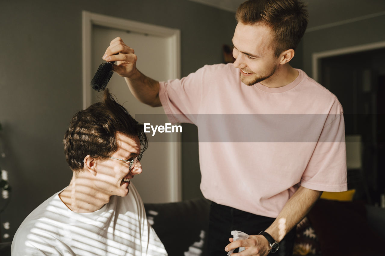 Young man combing male friend's hair while sitting at home