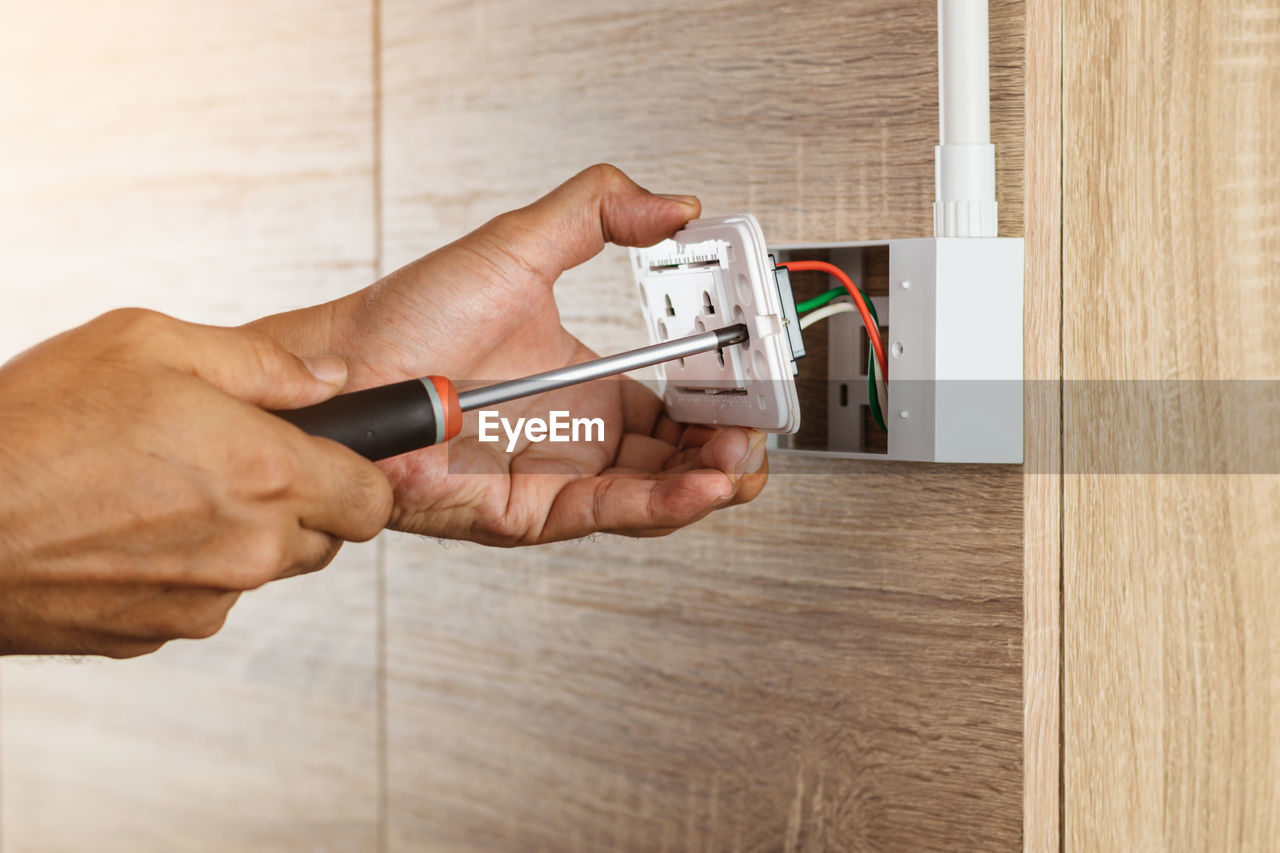 Electrician is using a screwdriver to install a power outlet in to a plastic box on a wooden wall.