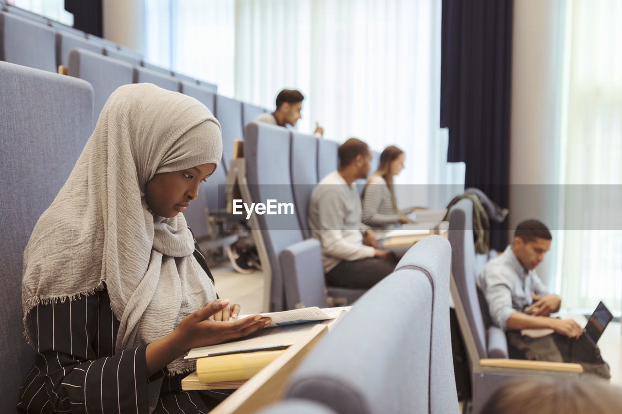 Serious woman in hijab reading book by students at university lecture hall