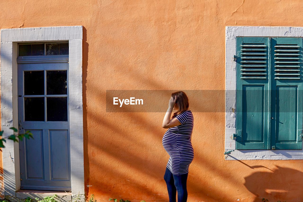 Side view of pregnant woman standing against wall of house