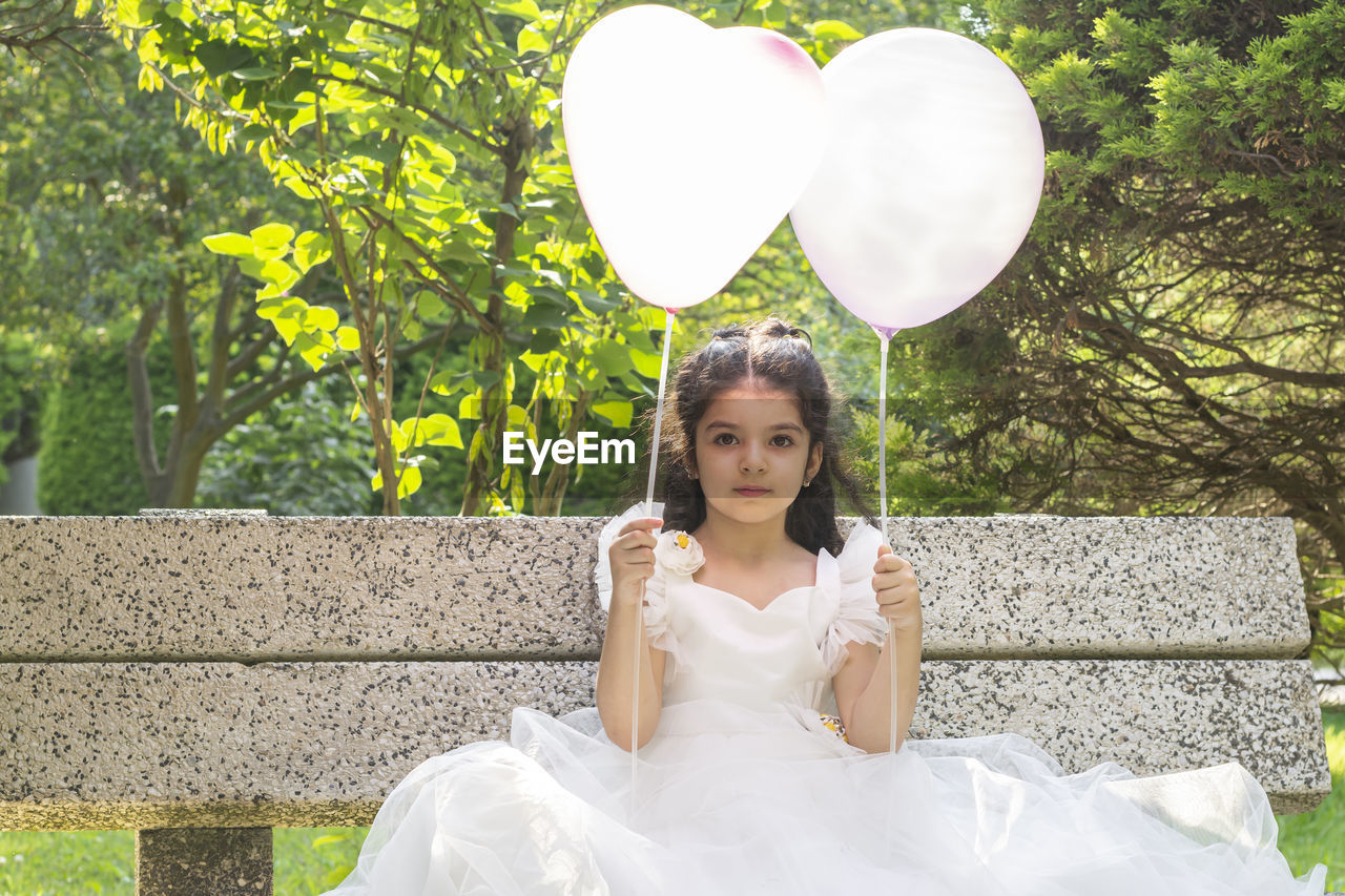 emotion, women, one person, wedding dress, positive emotion, portrait, plant, adult, love, wedding, young adult, bride, celebration, dress, event, happiness, female, nature, looking at camera, front view, smiling, white, sitting, balloon, tree, newlywed, heart shape, fashion, life events, brown hair, flower, outdoors, day, clothing, lifestyles, child, hairstyle, gown, ceremony, person, holding, long hair, bridal clothing, cheerful, contemplation, full length, looking, joy, leisure activity, childhood, romance