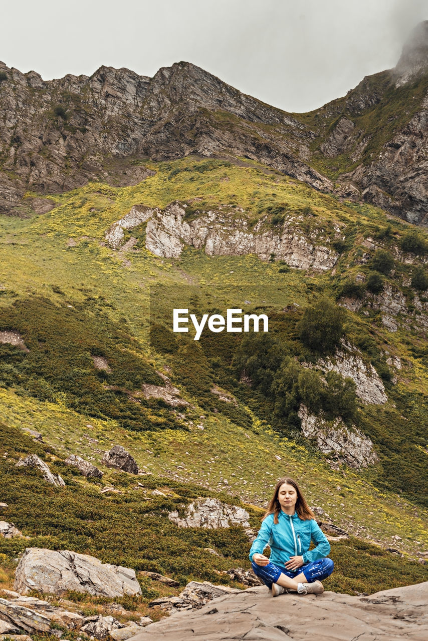 Meditating young woman in blue sitting on stone in  mountain valley,  consciousness, meditation