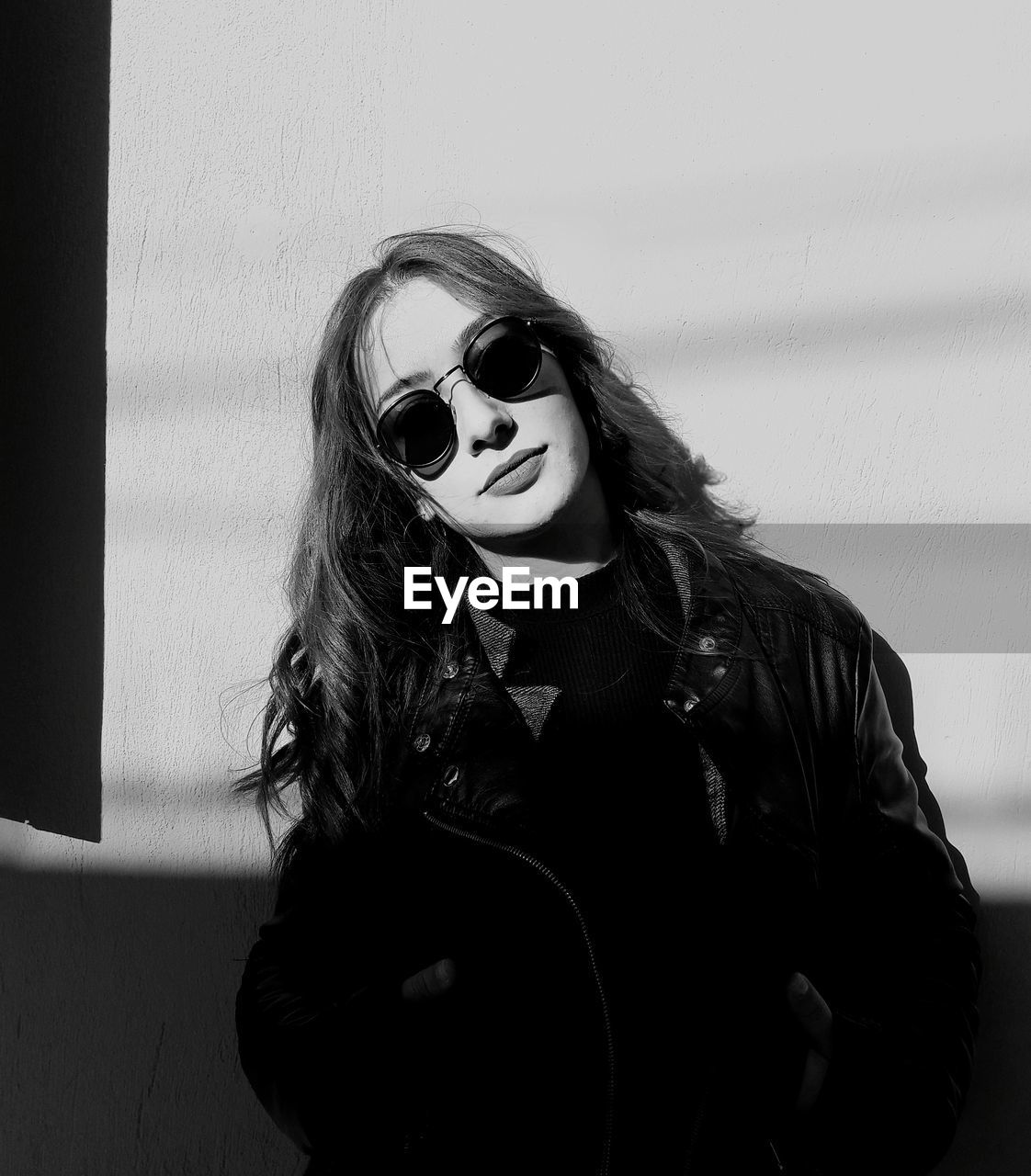 Portrait of young woman wearing sunglasses and jacket against wall