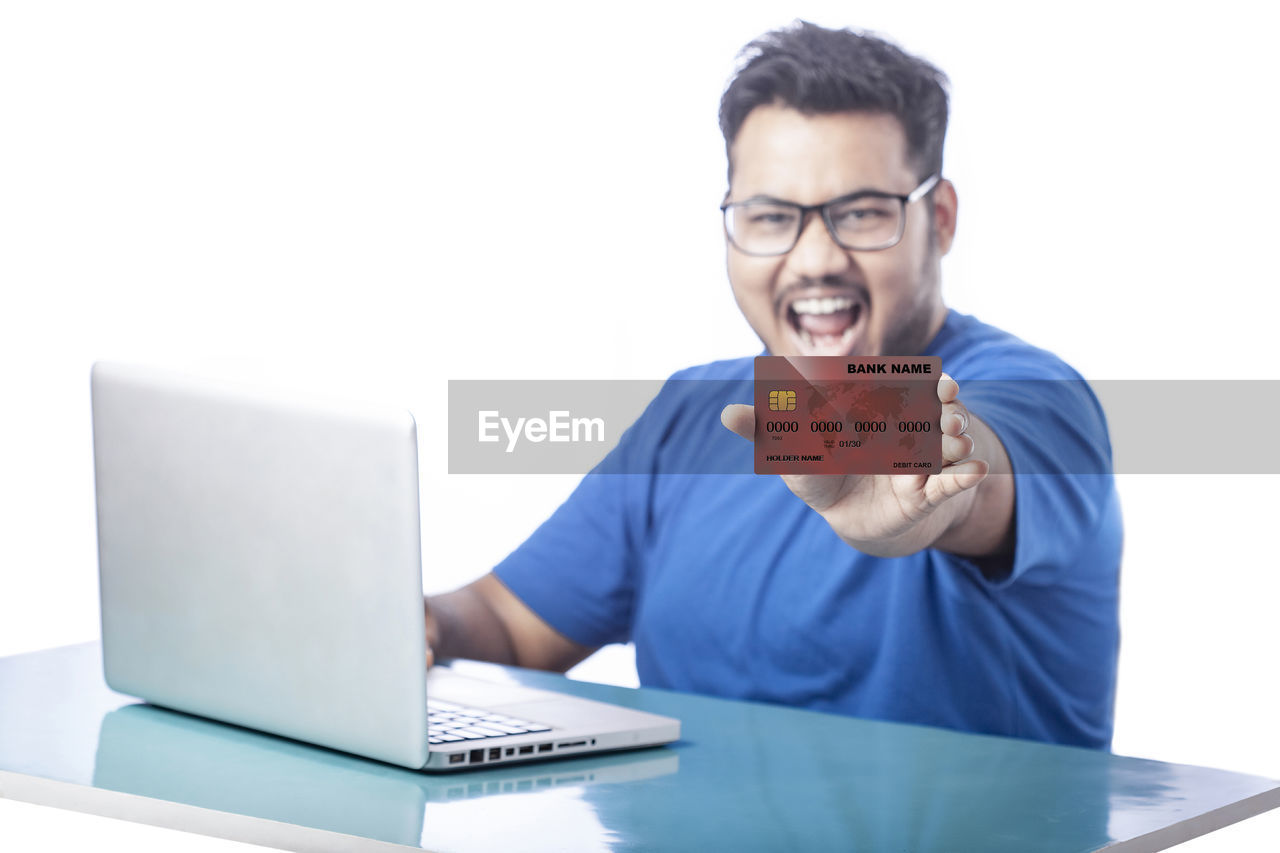 computer, one person, technology, laptop, adult, communication, wireless technology, business, men, eyeglasses, using laptop, glasses, smiling, indoors, writing, person, happiness, businessman, young adult, emotion, white background, portrait, looking, table, office, internet, casual clothing, occupation, cheerful, front view, desk, white-collar worker, computer network, computer equipment, furniture, working, studio shot, positive emotion, lifestyles, copy space, sitting
