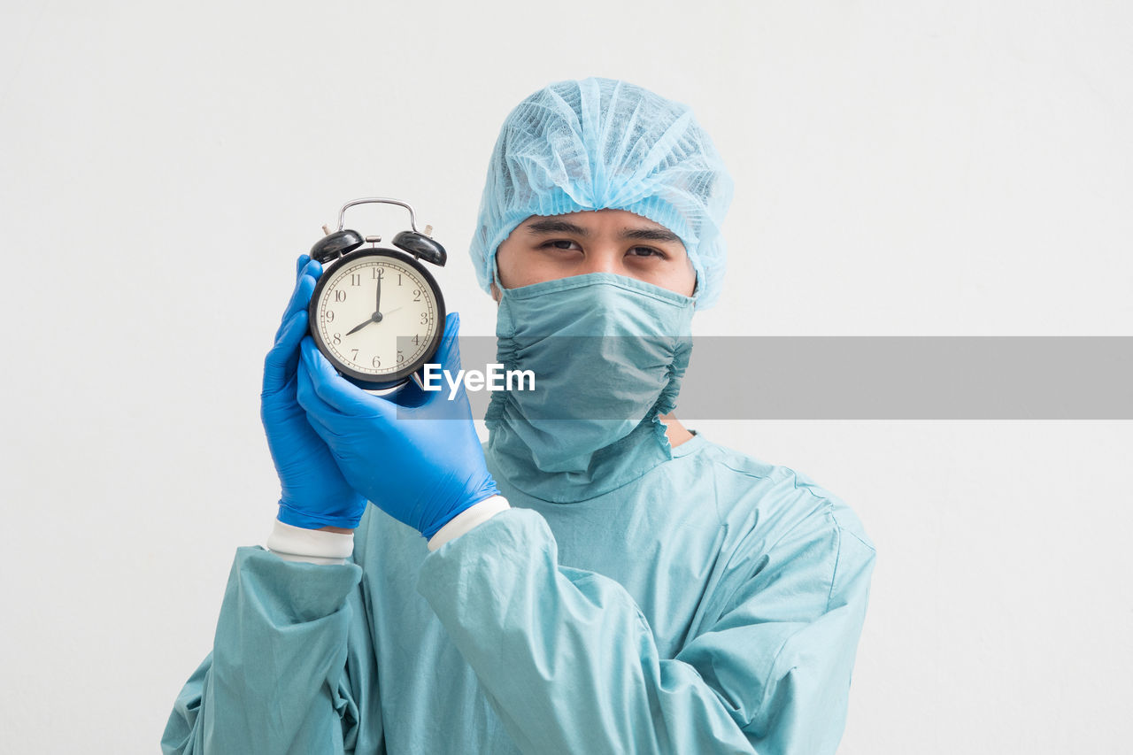 Portrait of surgeon holding alarm clock while standing against white background