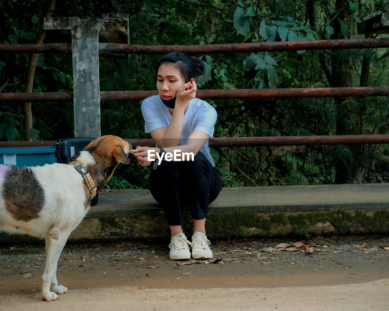 Full length of young woman with mask feeding dog outdoors