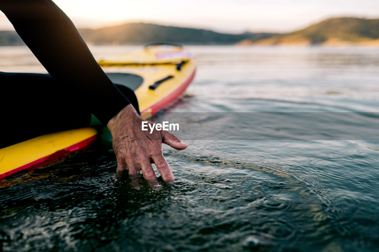 Crop unrecognizable male surfer sitting on sup board and touching sea water at sunset