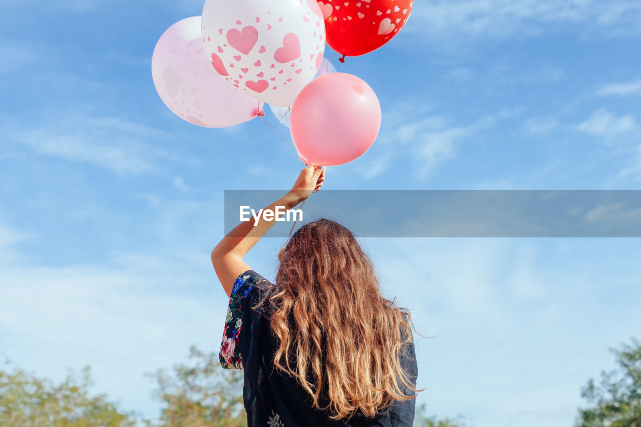 Rear view of woman holding helium balloons against blue sky