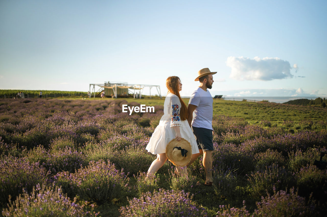 Side view of couple walking on lavender field against sky in sunny day