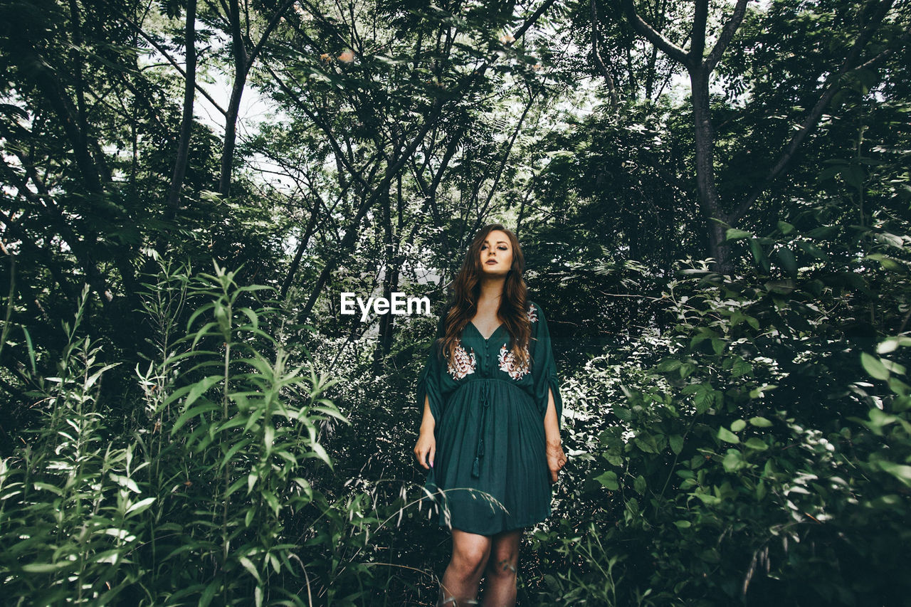 PORTRAIT OF YOUNG WOMAN STANDING BY PLANTS IN FOREST